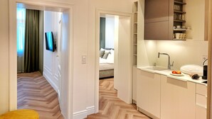 A modern hotel room with a herringbone wood floor, a small yellow ottoman, a flat-screen TV, a bed with white linens, and a kitchenette with a sink and countertop.