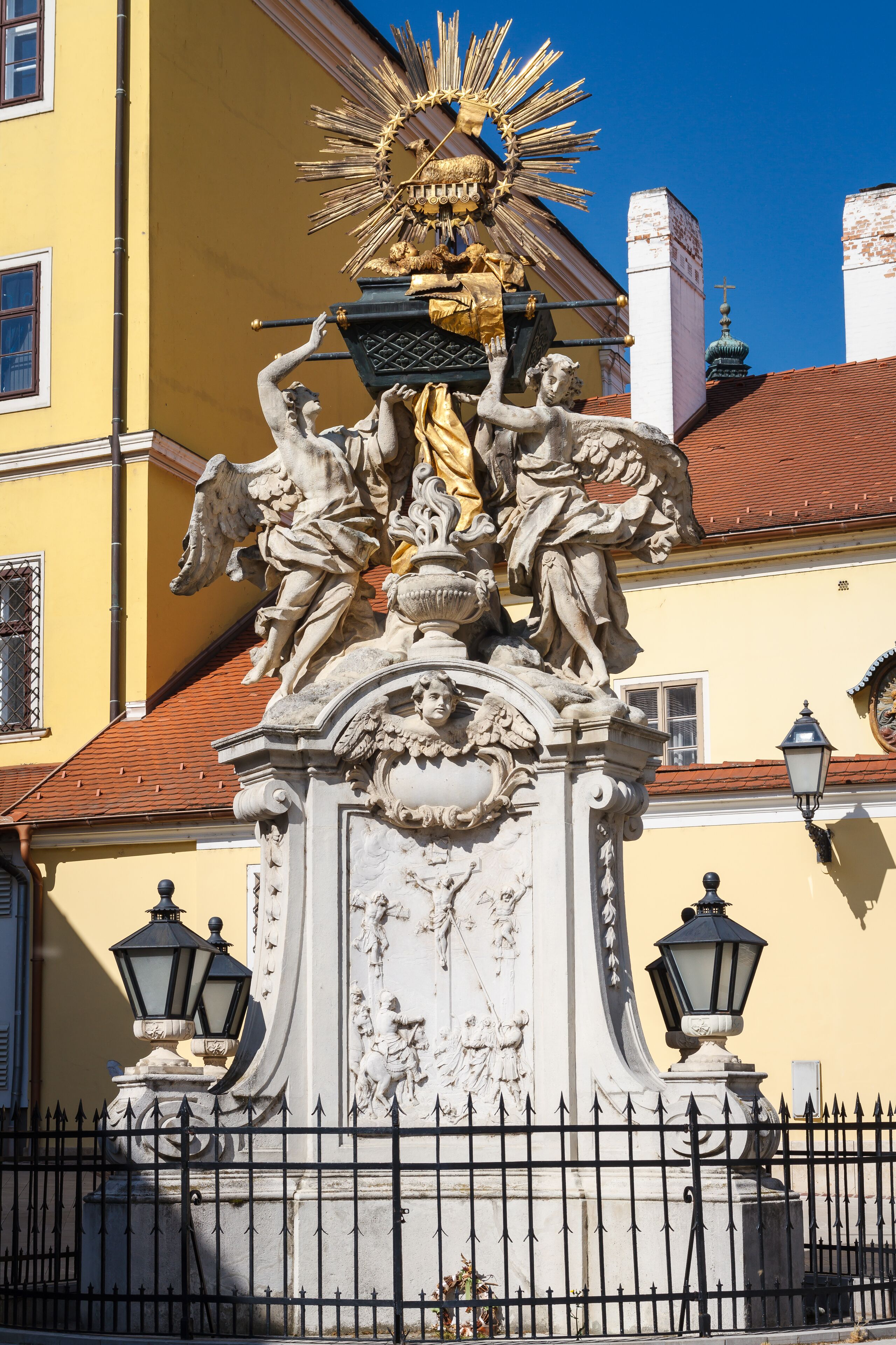 Une statue détaillée, surmontée d’une figure dorée, se dressant devant la façade jaune d’un immeuble et entourée d’une clôture en métal noir.