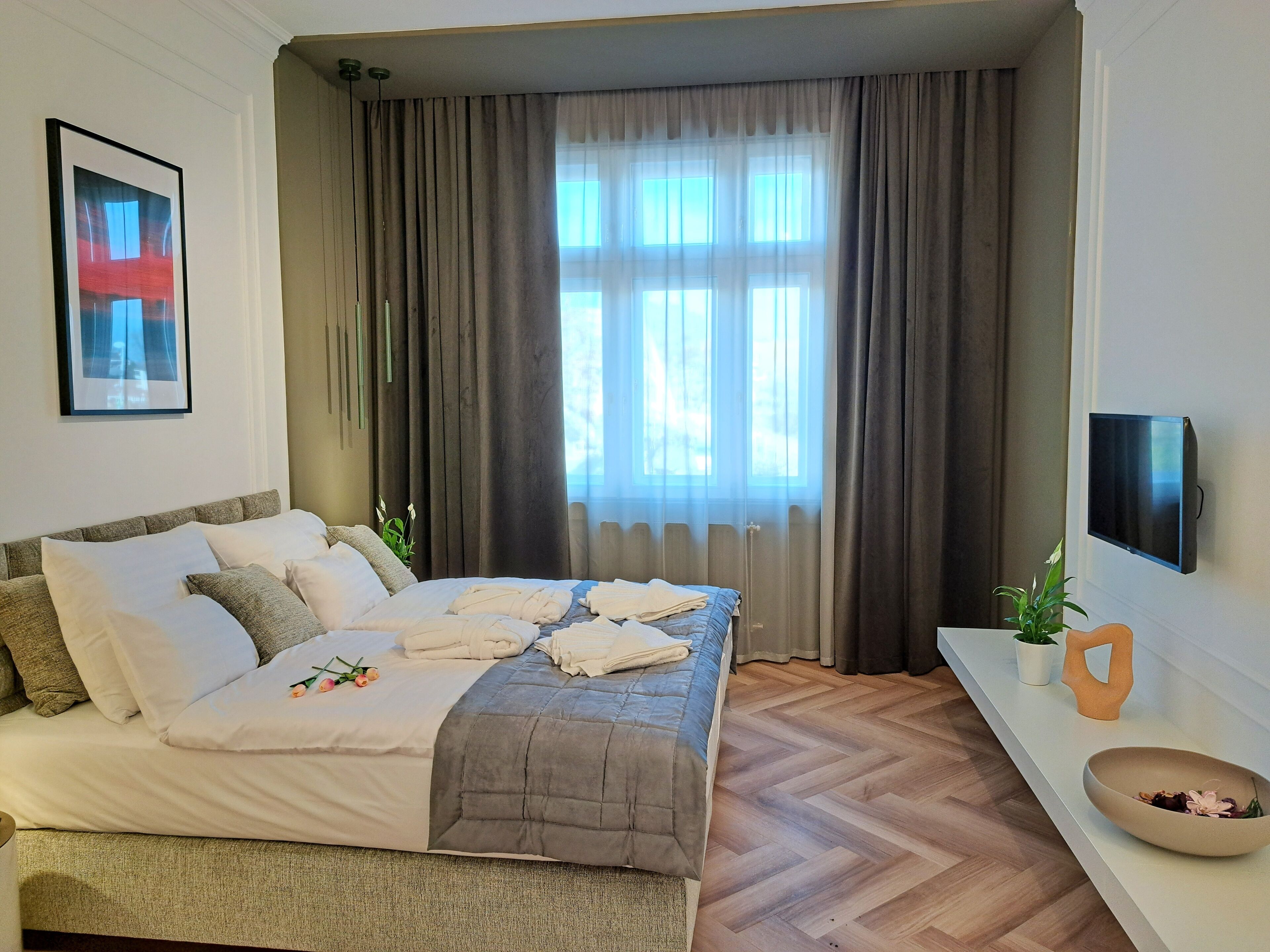 A modern hotel room with a large bed, a flat-screen TV mounted on the wall, and a window with curtains.