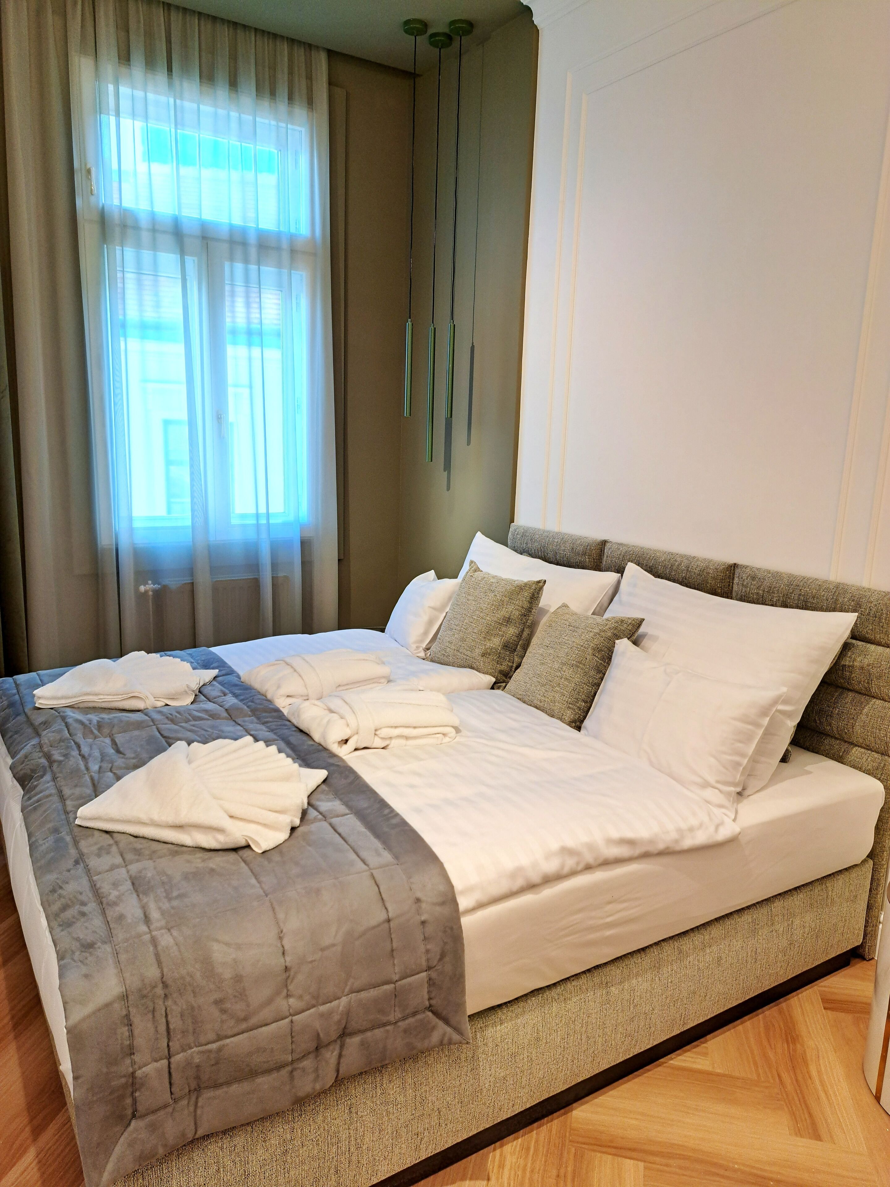 A neatly made bed with white linens and a gray quilted cover, a wooden headboard, and a window with sheer curtains allowing natural light.