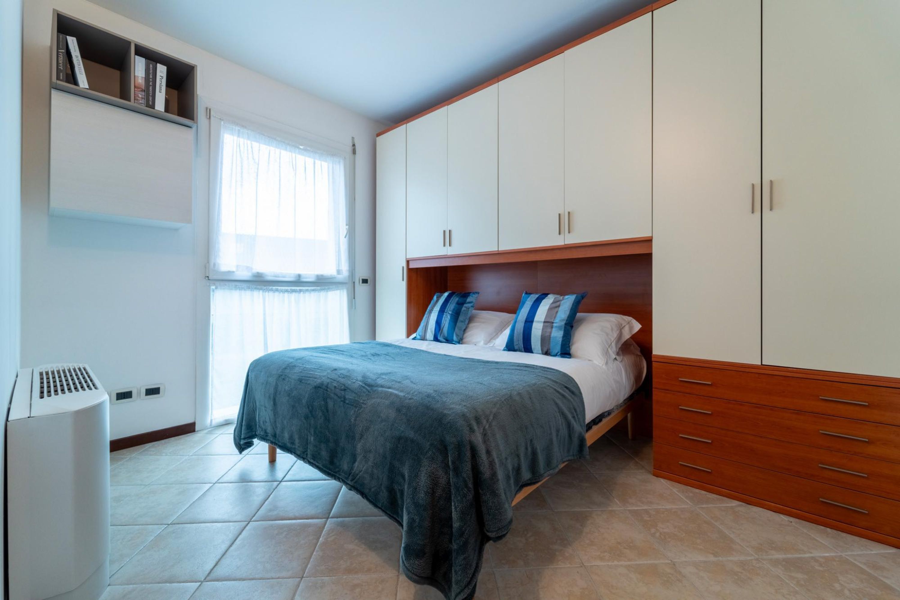 Un dormitorio con una cama, un armario, una estantería con libros y una ventana.