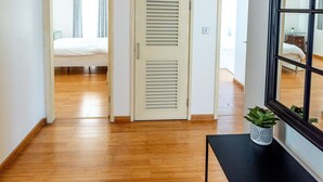 Un pasillo con suelo de madera, una cama al fondo, una puerta con paneles verticales y una consola negra con una planta.