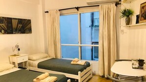 Hotellihuone, jossa on kaksi sänkyä, pieni yöpöytä lampun kanssa, ikkuna, jossa on verhot, pieni pyöreä pöytä ja ruukkukasvi.