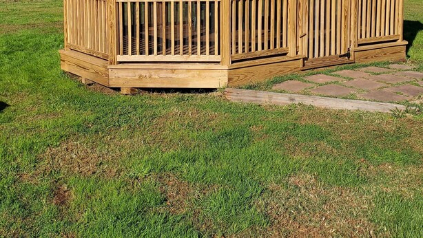 Un gazebo in legno con tetto a spiovente e lati aperti, immerso in un prato verdeggiante con alberi sullo sfondo.