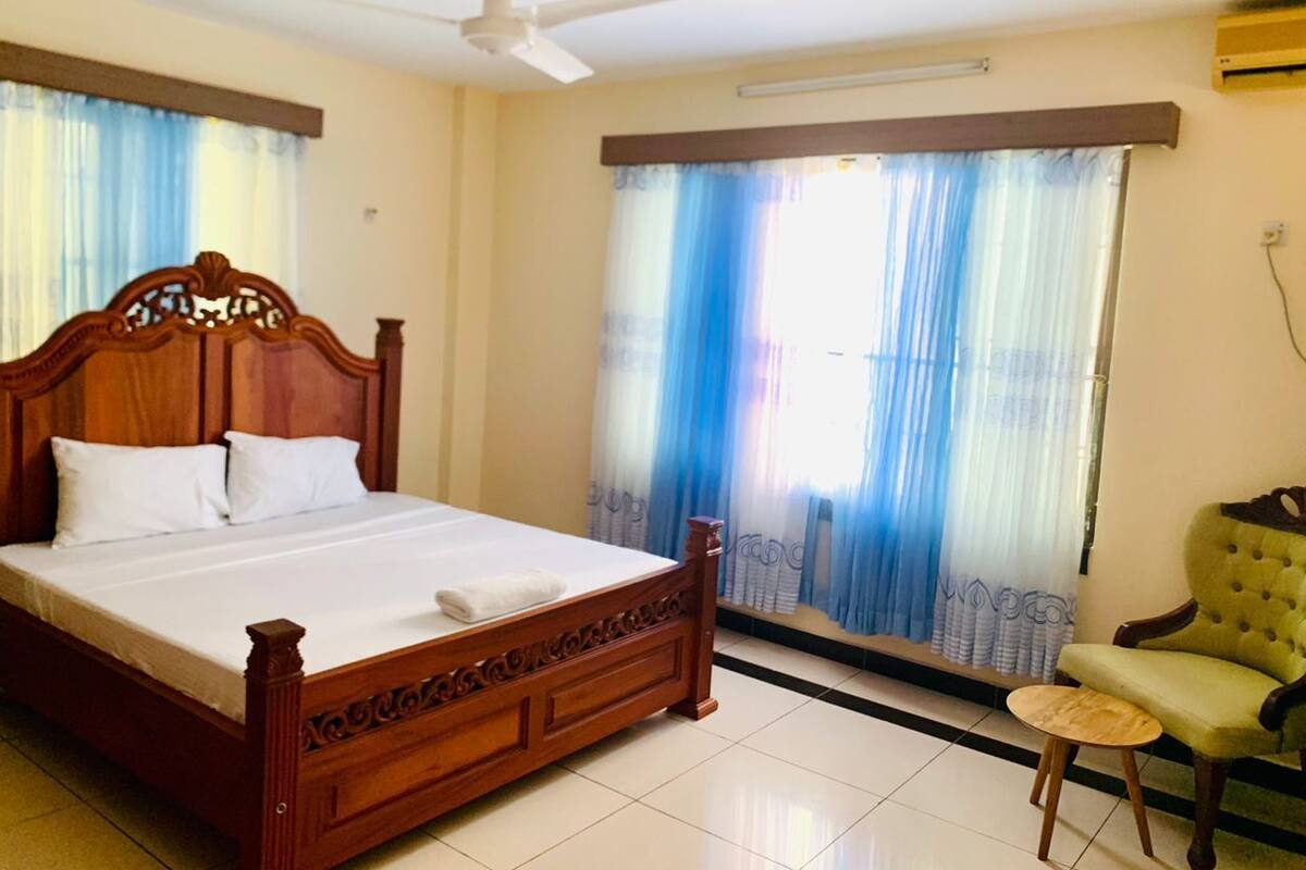 A bedroom with a wooden bed, a ceiling fan, a window with curtains, a small round table, and a green armchair.