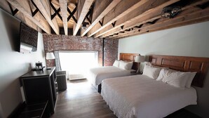 Habitación de hotel con pared de ladrillo, vigas de madera, dos camas, un escritorio y un televisor.