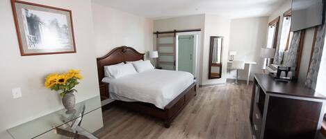 Ein Hotelzimmer mit einem hölzernen Bett, einem Glastisch mit Blumenvase, einem Schreibtisch mit Lampe und einem großen Spiegel.