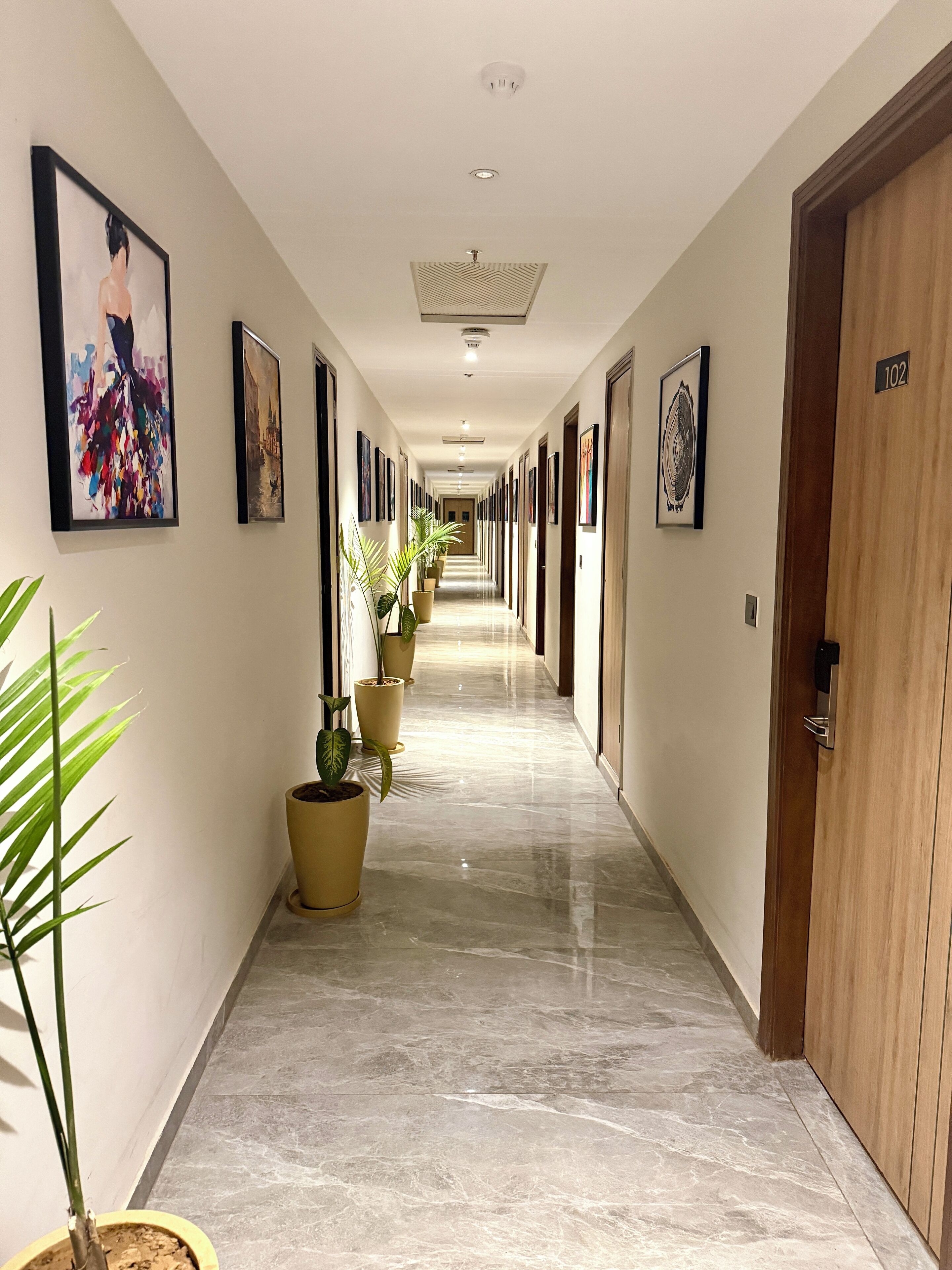 A long, well-lit hallway with multiple doors, potted plants, and framed artwork on the walls.
