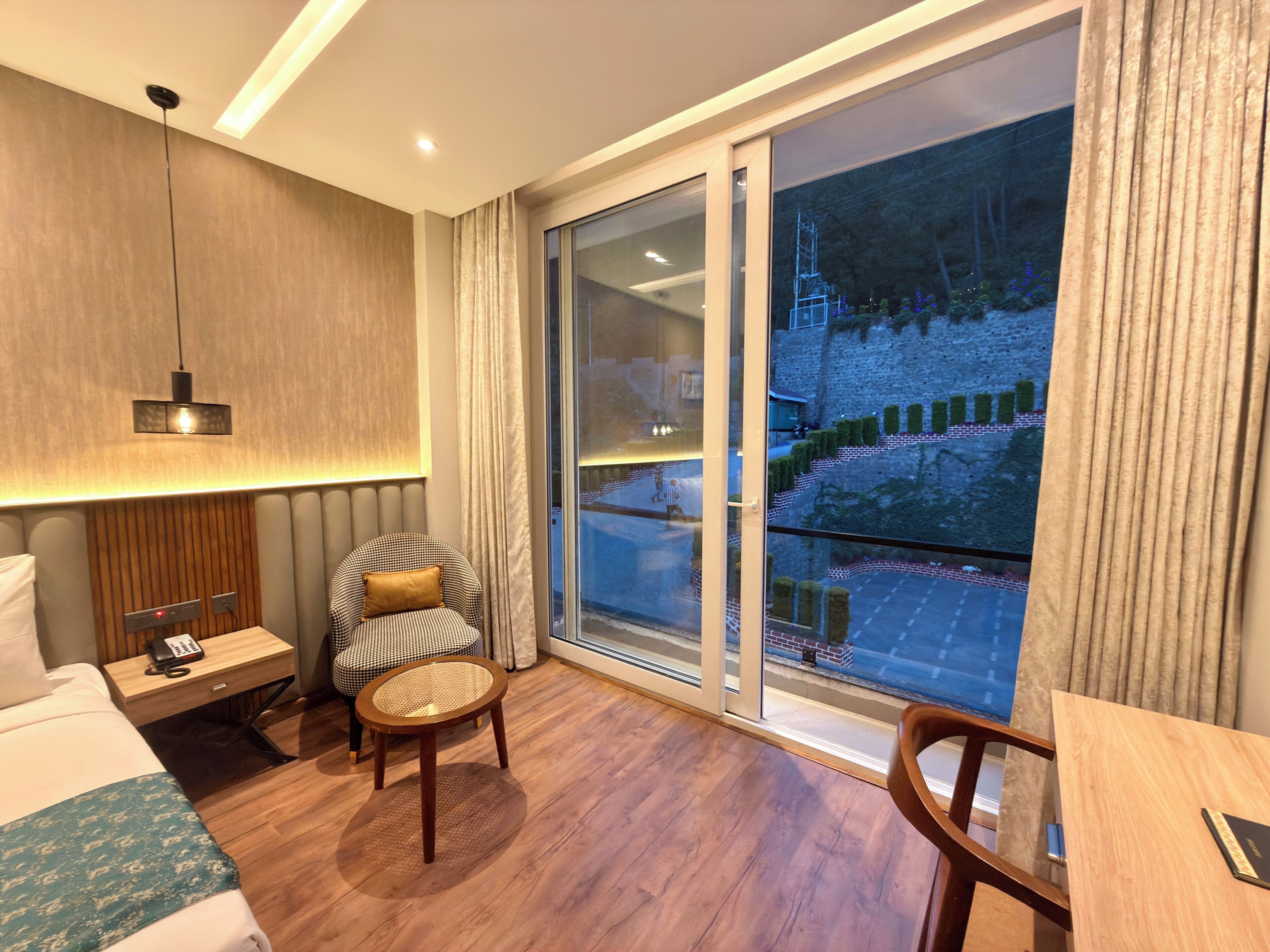 Habitación de hotel con cama, una silla, una mesita pequeña, una mesita de noche con teléfono, una lámpara y un amplio ventanal con vistas a un edificio y una calle.