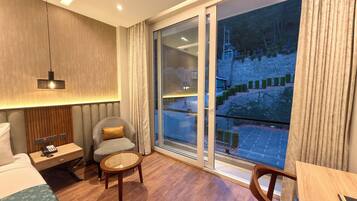 Habitación de hotel con cama, una silla, una mesita pequeña, una mesita de noche con teléfono, una lámpara y un amplio ventanal con vistas a un edificio y una calle.