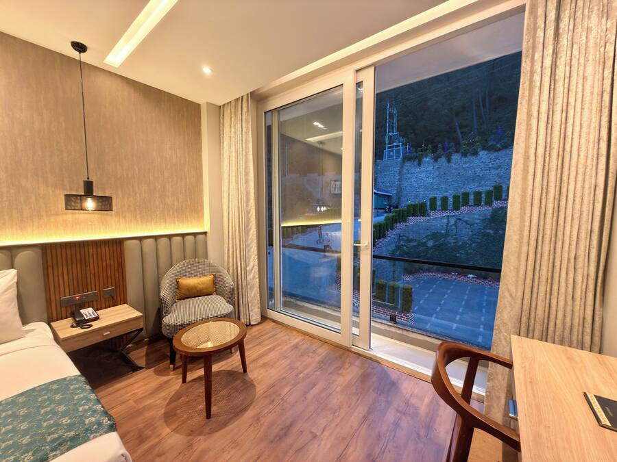 Habitación de hotel con cama, una silla, una mesita pequeña, una mesita de noche con teléfono, una lámpara y un amplio ventanal con vistas a un edificio y una calle.