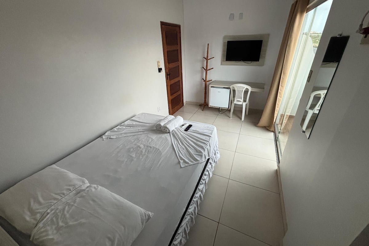 A small, clean room with a bed, a desk, a chair, a TV, and a window with curtains.