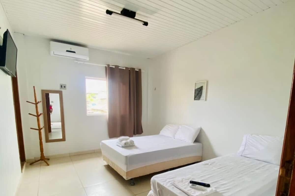 A room with two beds, a TV, an air conditioner, a window with curtains, and a wooden coat rack.