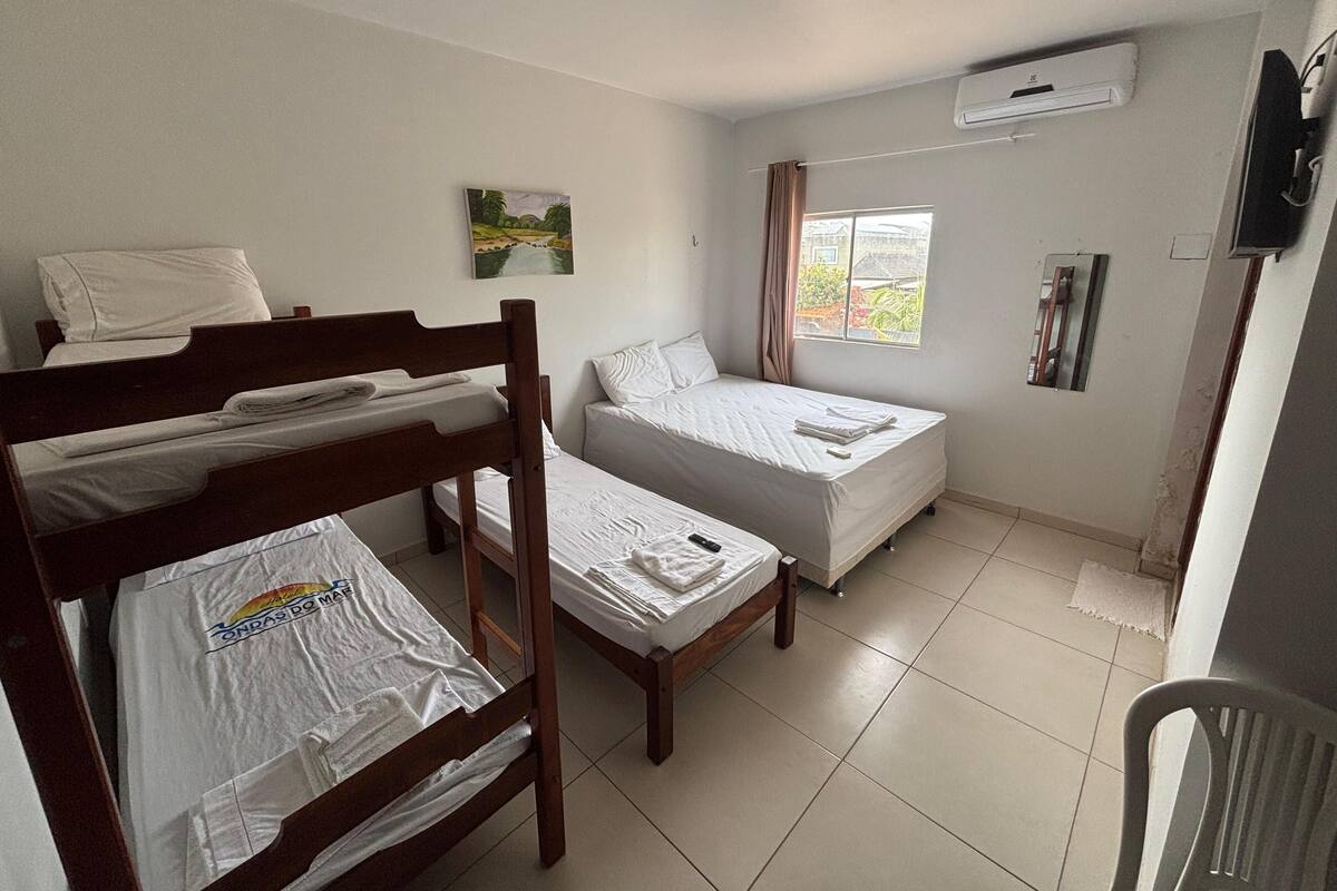 A room with two single beds, a bunk bed, a window with curtains, an air conditioning unit, and a television.