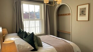 A bedroom with a bed, two bedside lamps, a window with curtains, a chandelier, a framed picture, and a built-in shelf.