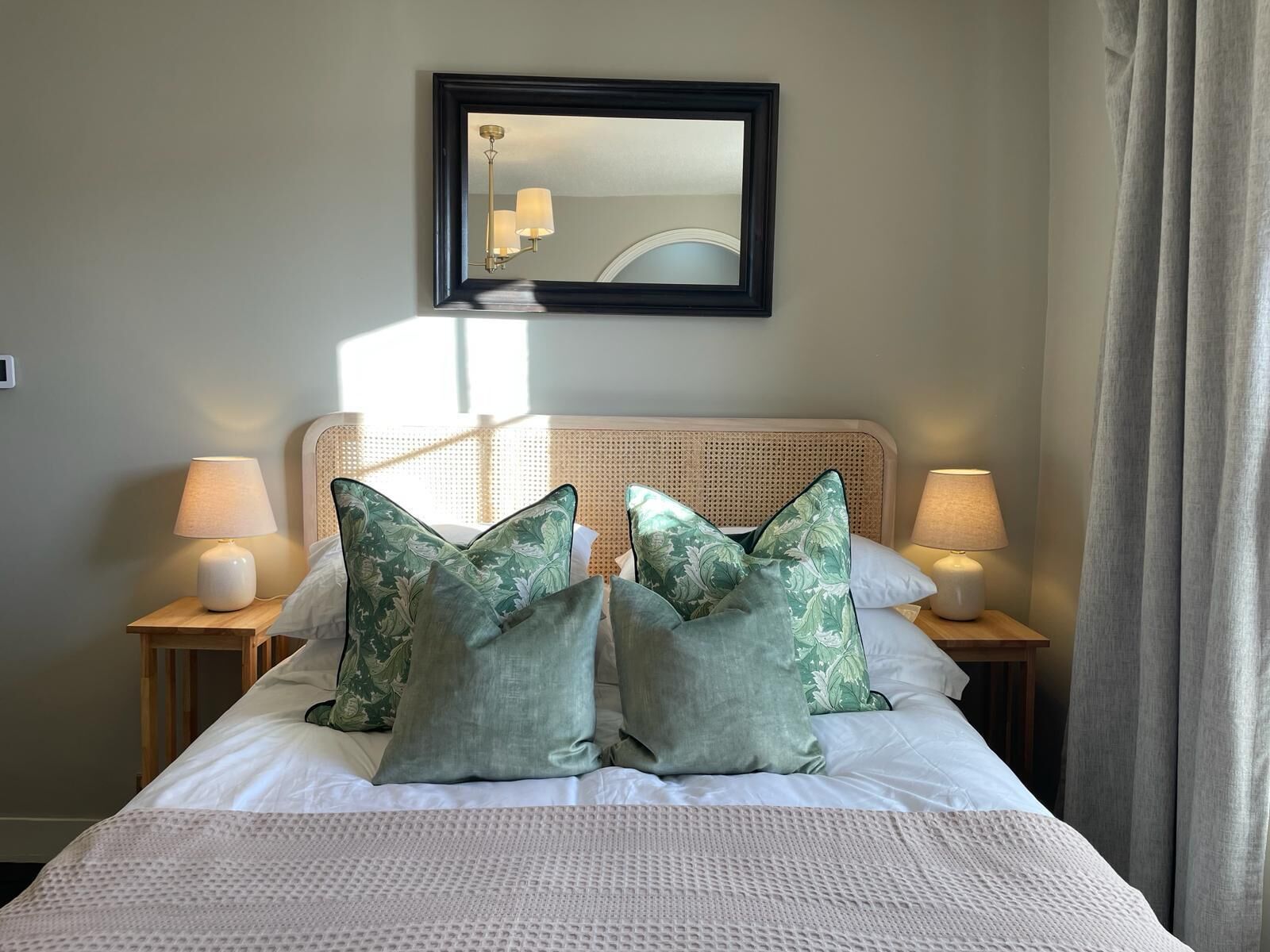 Una cama bien hecha con edredón blanco, dos almohadas florales verdes y dos lámparas de noche sobre mesitas de madera.