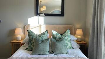 Una cama bien hecha con edredón blanco, dos almohadas florales verdes y dos lámparas de noche sobre mesitas de madera.