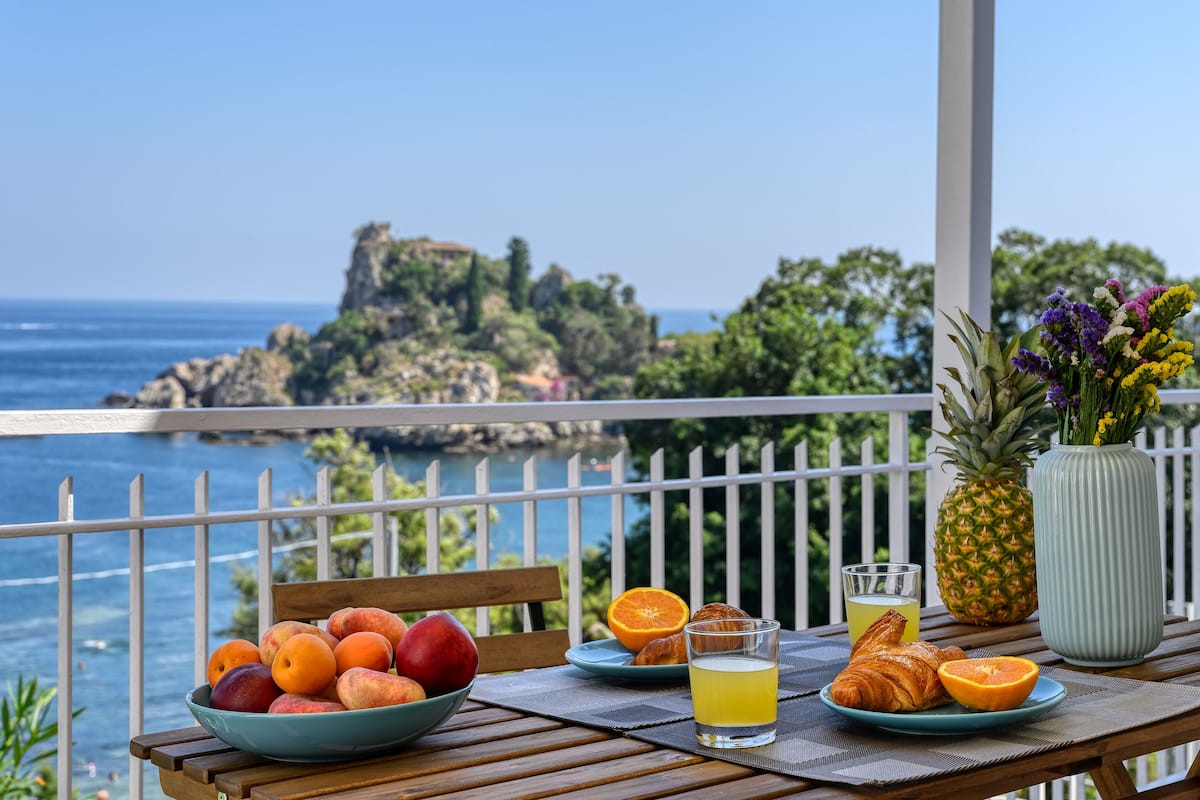 A table with fruits, a pineapple, a glass of juice, and a croissant on a balcony with a view of the ocean and rocky cliffs.