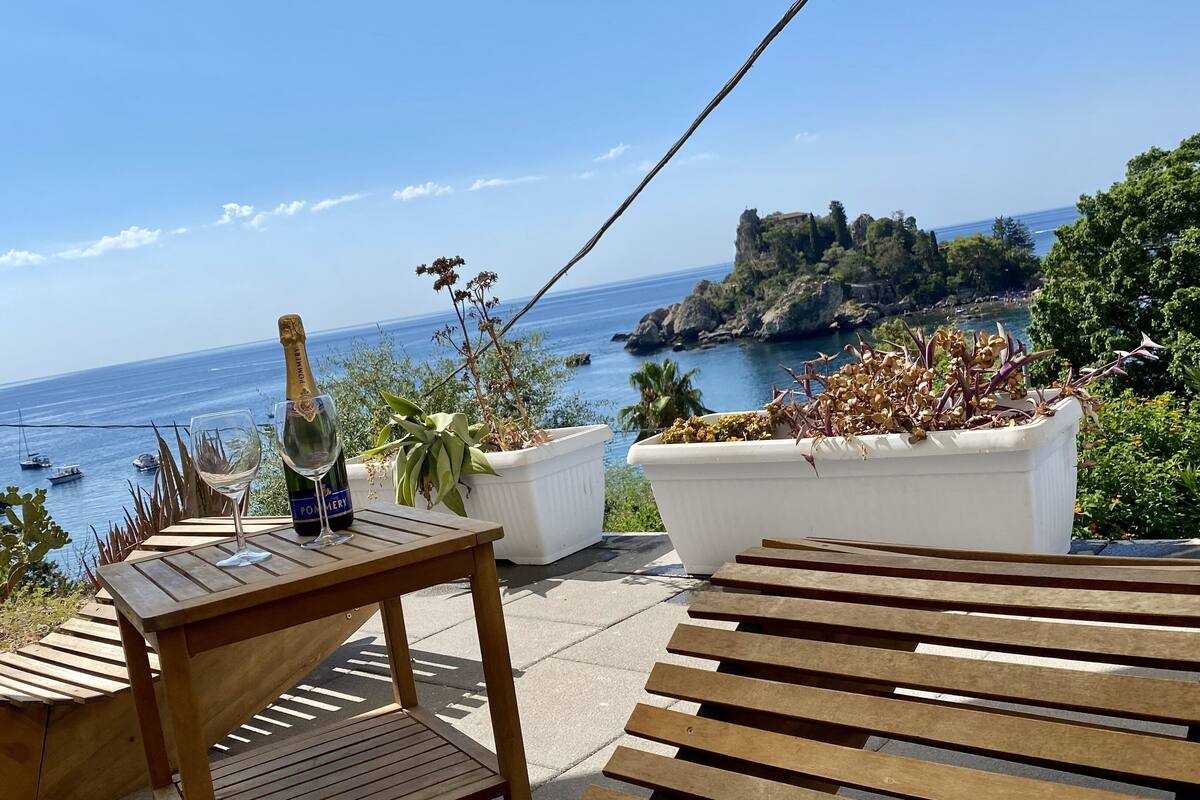 A wooden table with a bottle of champagne and a glass, overlooking a coastal view with boats and rocky cliffs.