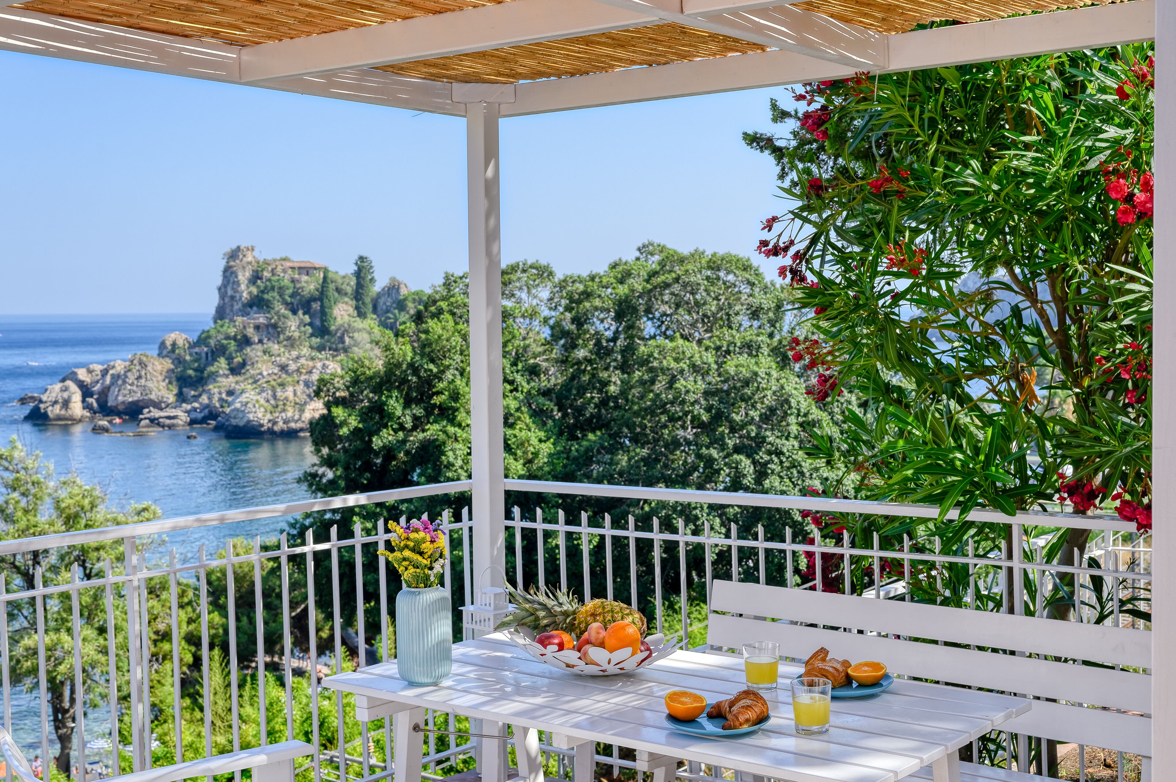 A balcony with a table set for breakfast, overlooking a coastal landscape with a rocky outcrop and trees.