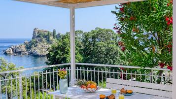 A balcony with a table set for breakfast, overlooking a coastal landscape with a rocky outcrop and trees.