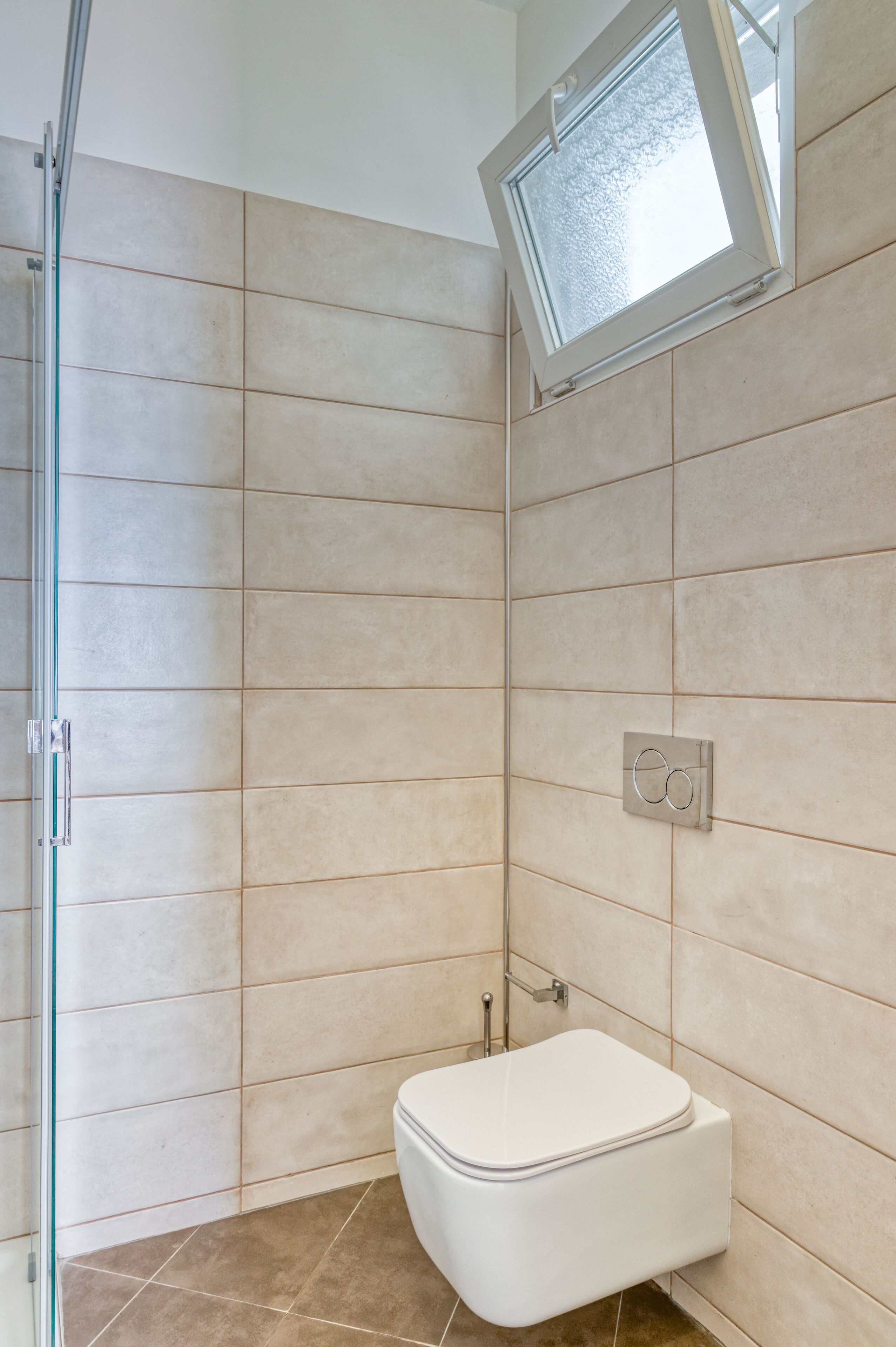 Un baño con azulejos color beige, un inodoro blanco y una ventana.