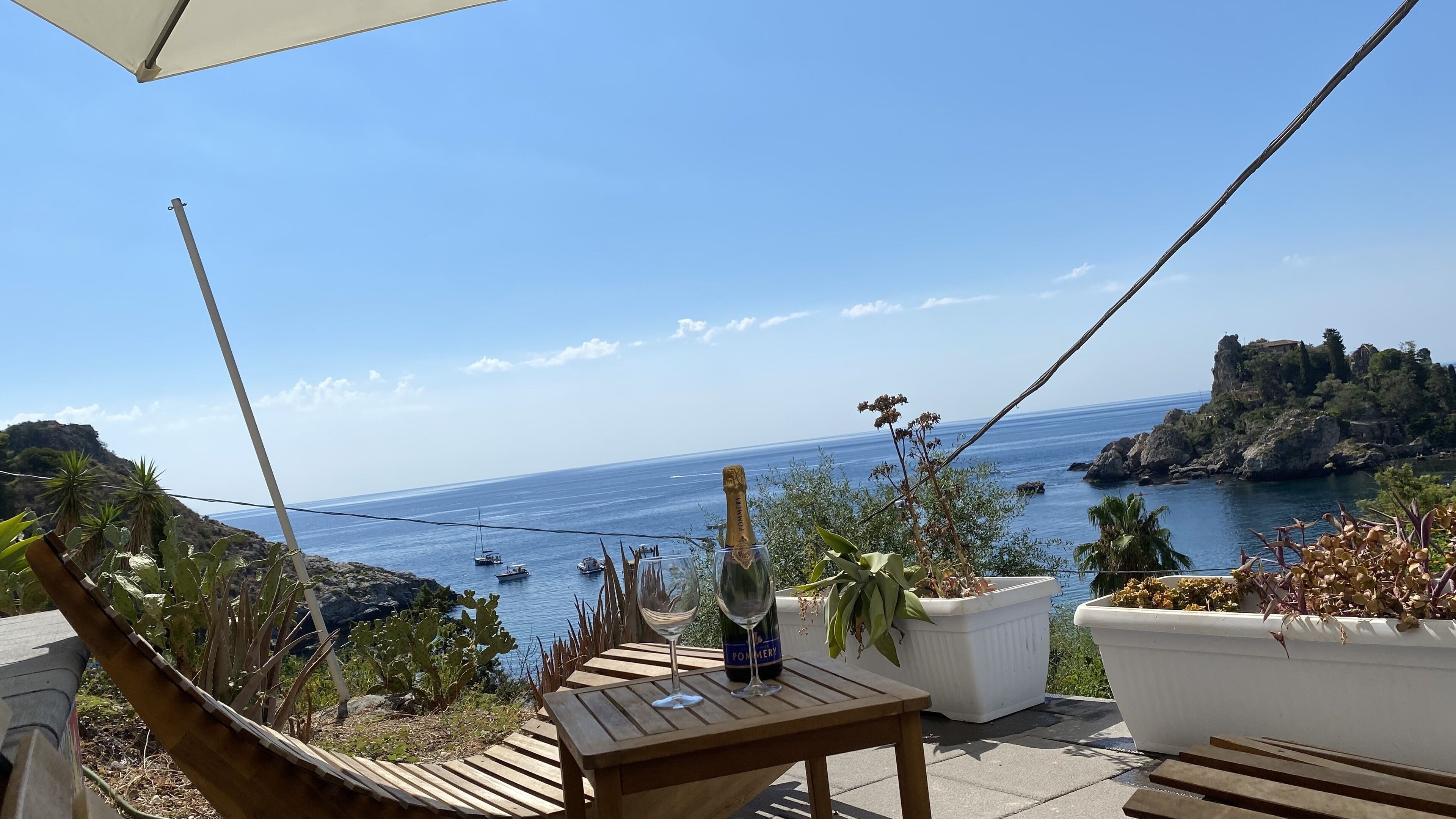 Un tavolo in legno con una bottiglia di champagne e un bicchiere, con vista sulla costa, barche e scogliere rocciose.