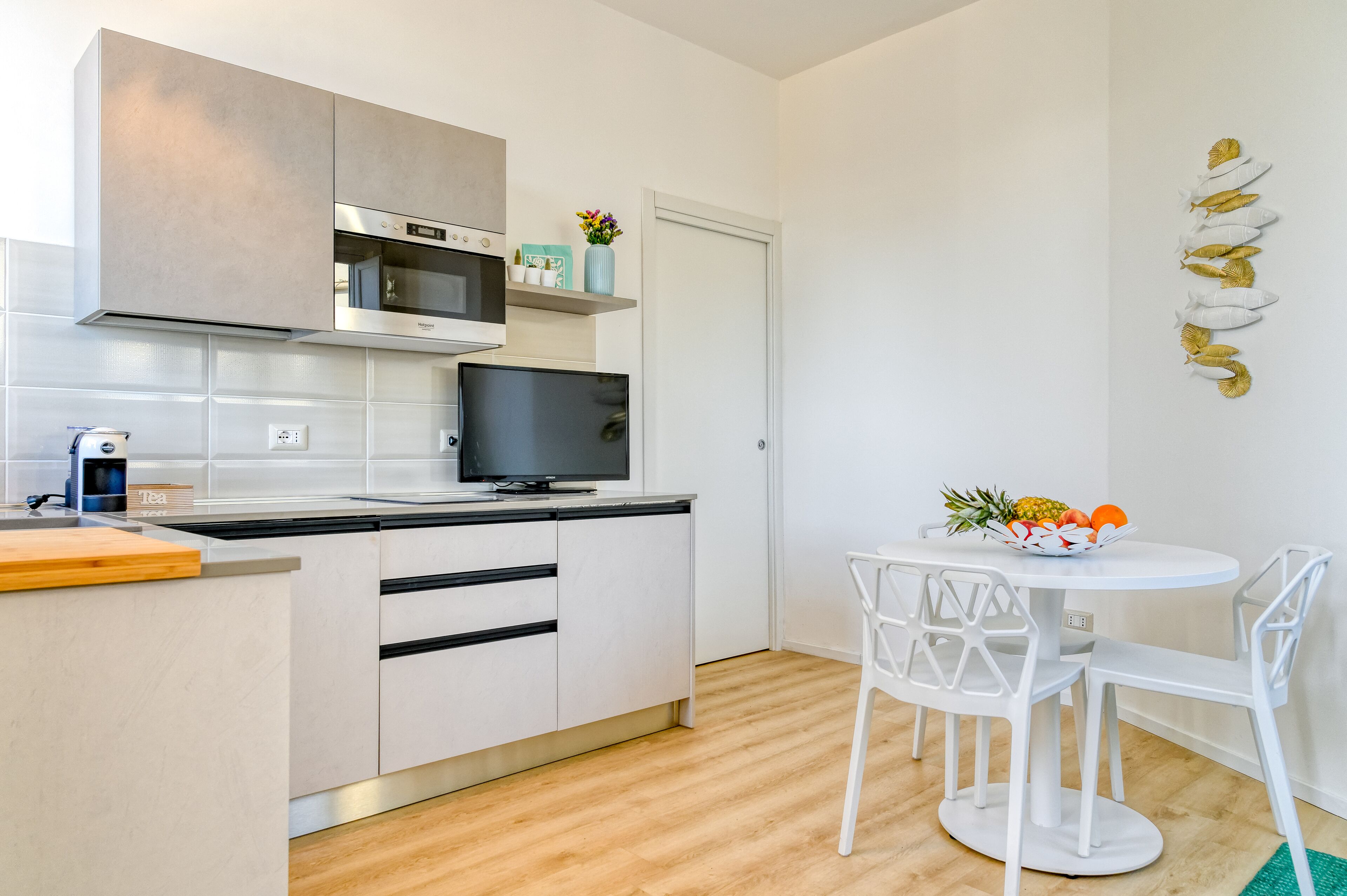 Una cucina moderna con mobili bianchi, un forno a microonde e una TV. C'è un'area pranzo con un tavolo e sedie bianche, e una ciotola di frutta sul tavolo.