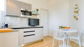 Una cucina moderna con mobili bianchi, un forno a microonde e una TV. C'è un'area pranzo con un tavolo e sedie bianche, e una ciotola di frutta sul tavolo.