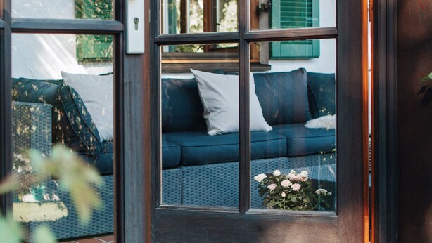 Une porte en bois avec des panneaux de verre qui donne sur une pièce où se trouvent un canapé bleu et un panier de fleurs.