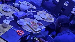 Una habitación con una televisión mostrando un canal de noticias, una mesa de comedor con platos, tazas y cubiertos, y una persona sosteniendo un teléfono inteligente.