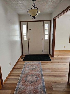 Un couloir doté d’un plancher en bois, d’un tapis à motifs, d’une porte avec un paillasson et d’un luminaire au plafond.
