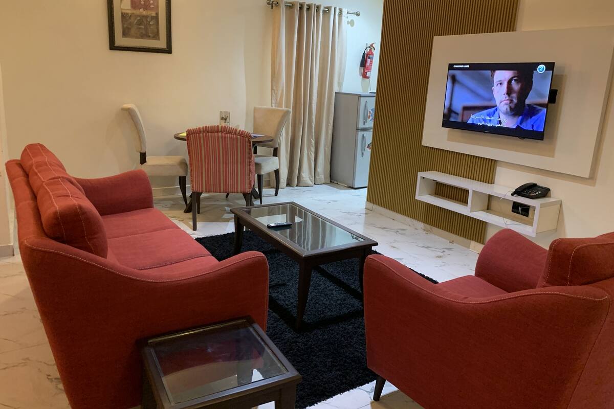 A living room with red sofas, a glass coffee table, a TV on the wall, a small table, and a chair.