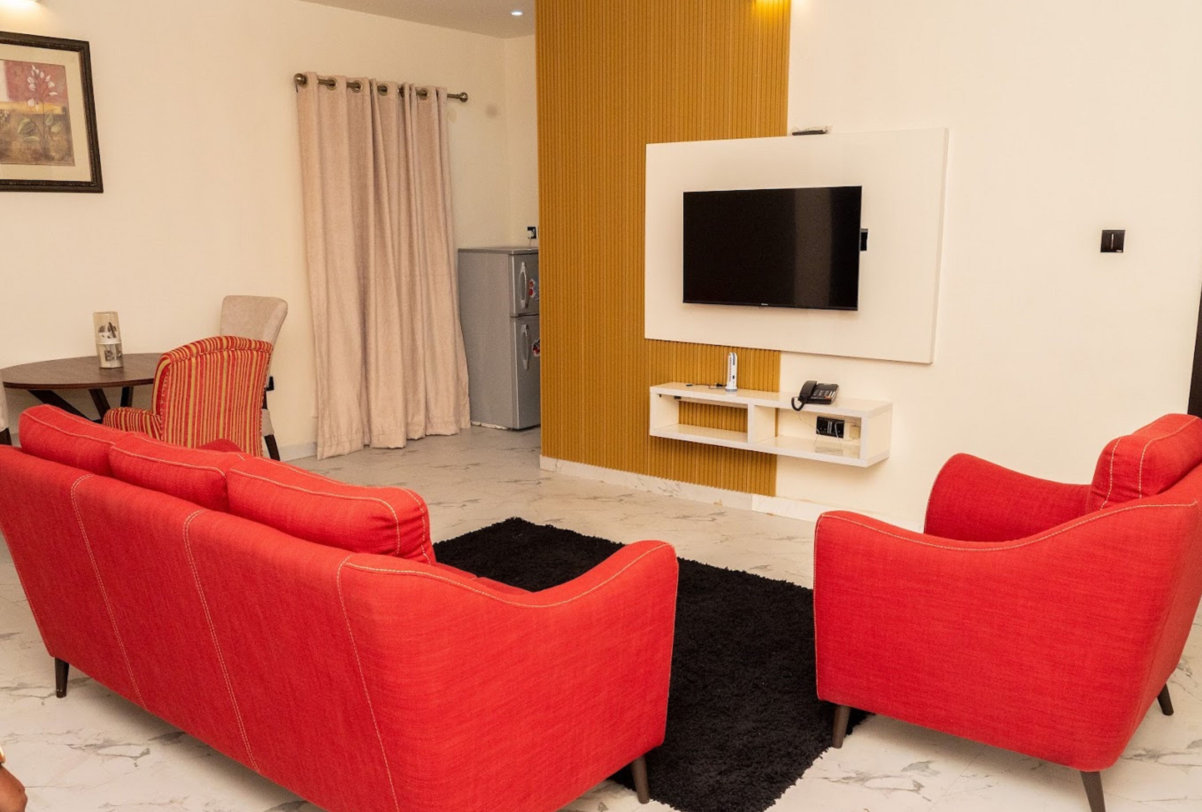 Una sala de estar con sofás rojos, un televisor montado en la pared, una mesita y un refrigerador.