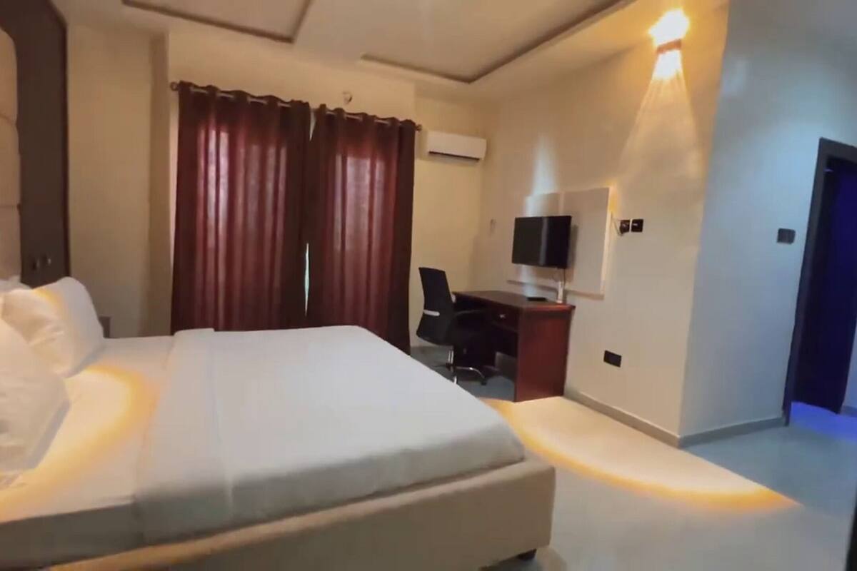 A hotel room with a large bed, a desk with a chair, a television, and an air conditioning unit.
