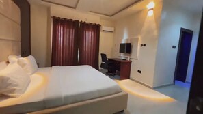 A hotel room with a large bed, a desk with a chair, a television, and an air conditioning unit. - Old English Torf Apartment (Owerri)