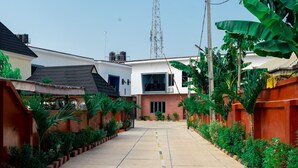 A narrow, paved pathway flanked by lush greenery and residential buildings with distinct architectural styles. - Old English Torf Apartment (Owerri)