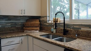 A kitchen with a granite countertop, a stainless steel sink, white cabinets, and a window with a view of trees. - Large 4bd 2bth pool home with hot tub, waterfall feature and private back yard! (Palm Bay)