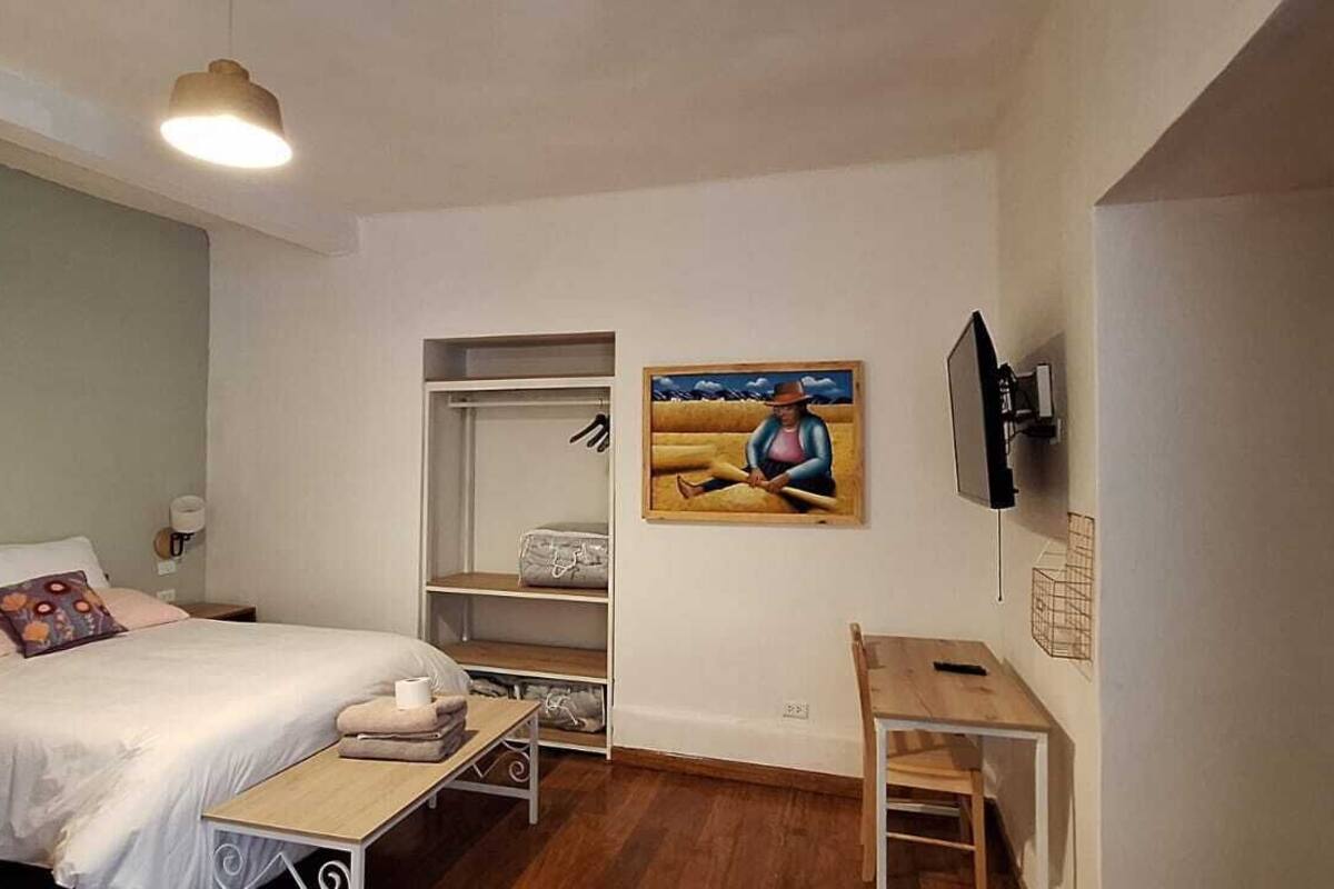 A bedroom with a bed, bedside table, and a desk. There is a wardrobe and a painting on the wall.