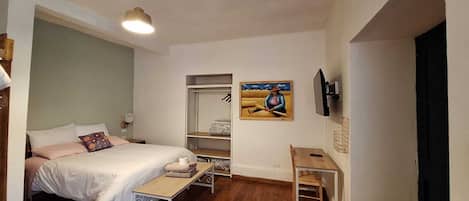 Quarto com cama, mesa de cabeceira e escrivaninha. Há um guarda-roupa e um quadro na parede.