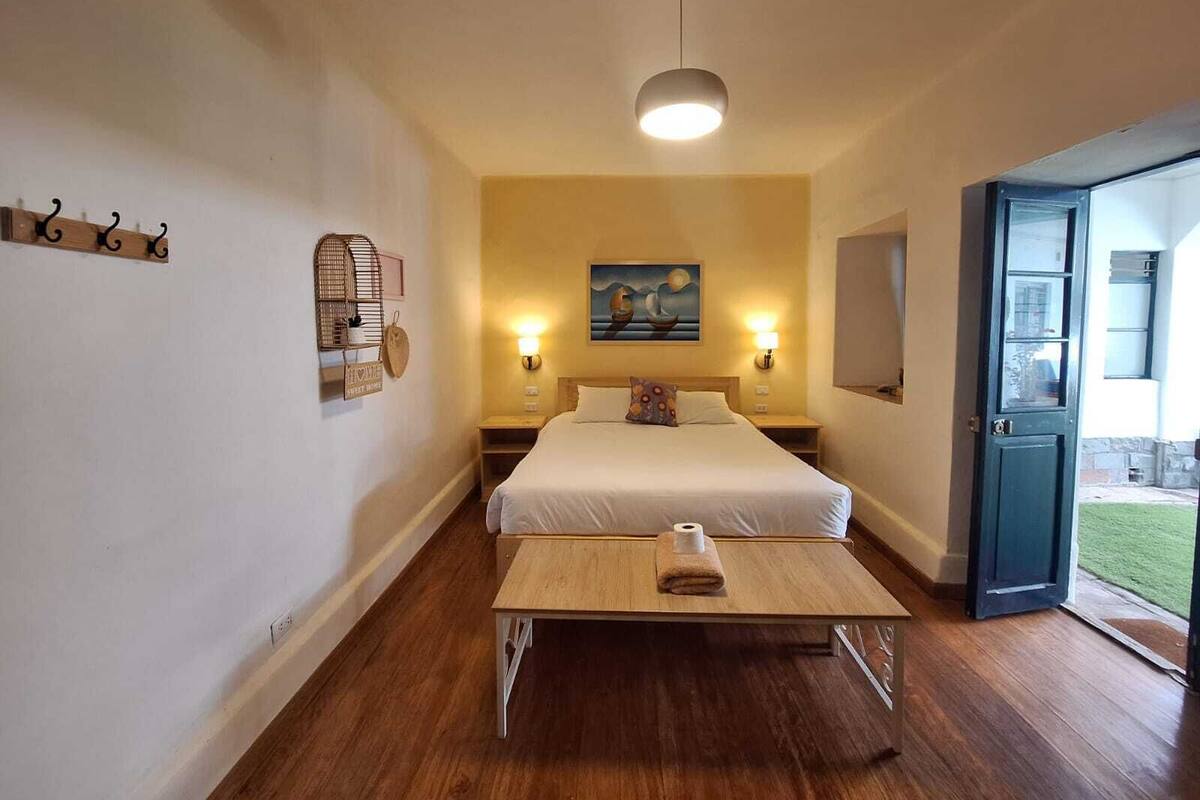 A hotel room with a bed, bedside tables, a small table with a towel, wall-mounted lights, a painting, and a door leading outside.