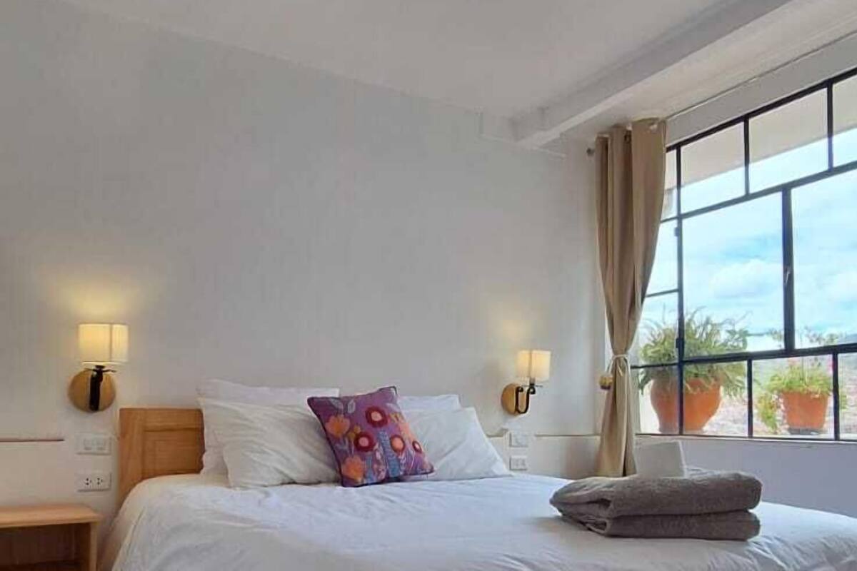 A bedroom with a large bed, bedside lamps, a wooden nightstand, and a view of the outdoors through a window.