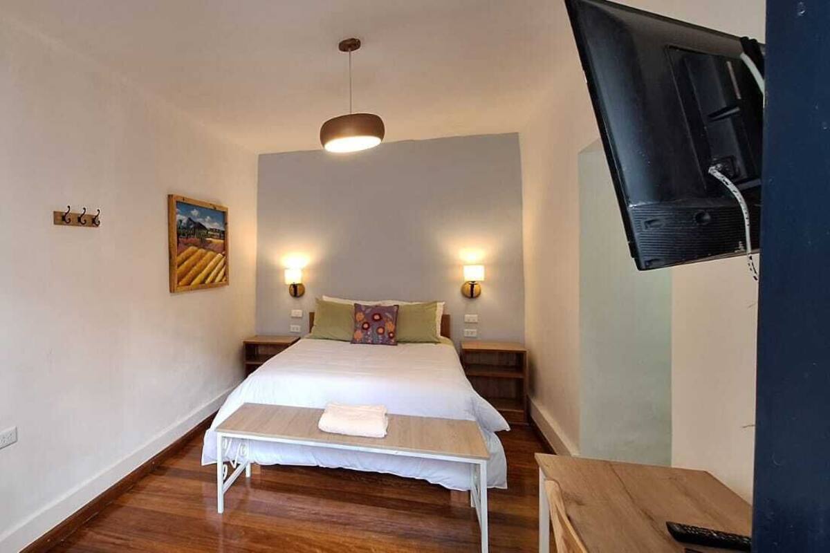 A bedroom with a bed, bedside tables, a hanging coat rack, a painting on the wall, and a television mounted on the wall.
