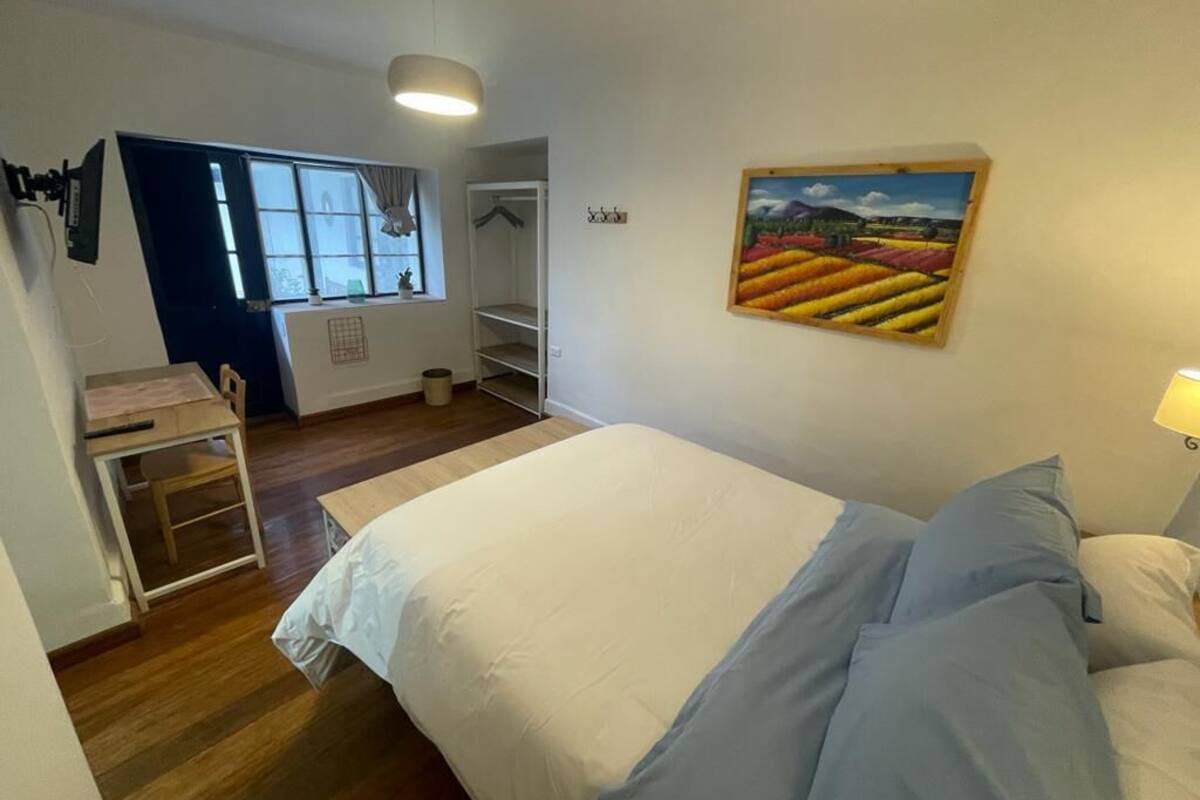A bedroom with a bed, a desk, and a chair. There is a window with curtains, a painting on the wall, and a lamp on the nightstand.