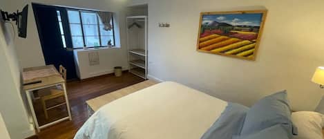 A bedroom with a bed, a desk, and a chair. There is a window with curtains, a painting on the wall, and a lamp on the nightstand.