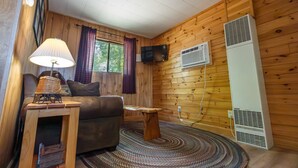 Una habitación acogedora con paredes de madera, un sofá, una mesa con lámpara, un televisor y un aire acondicionado.
