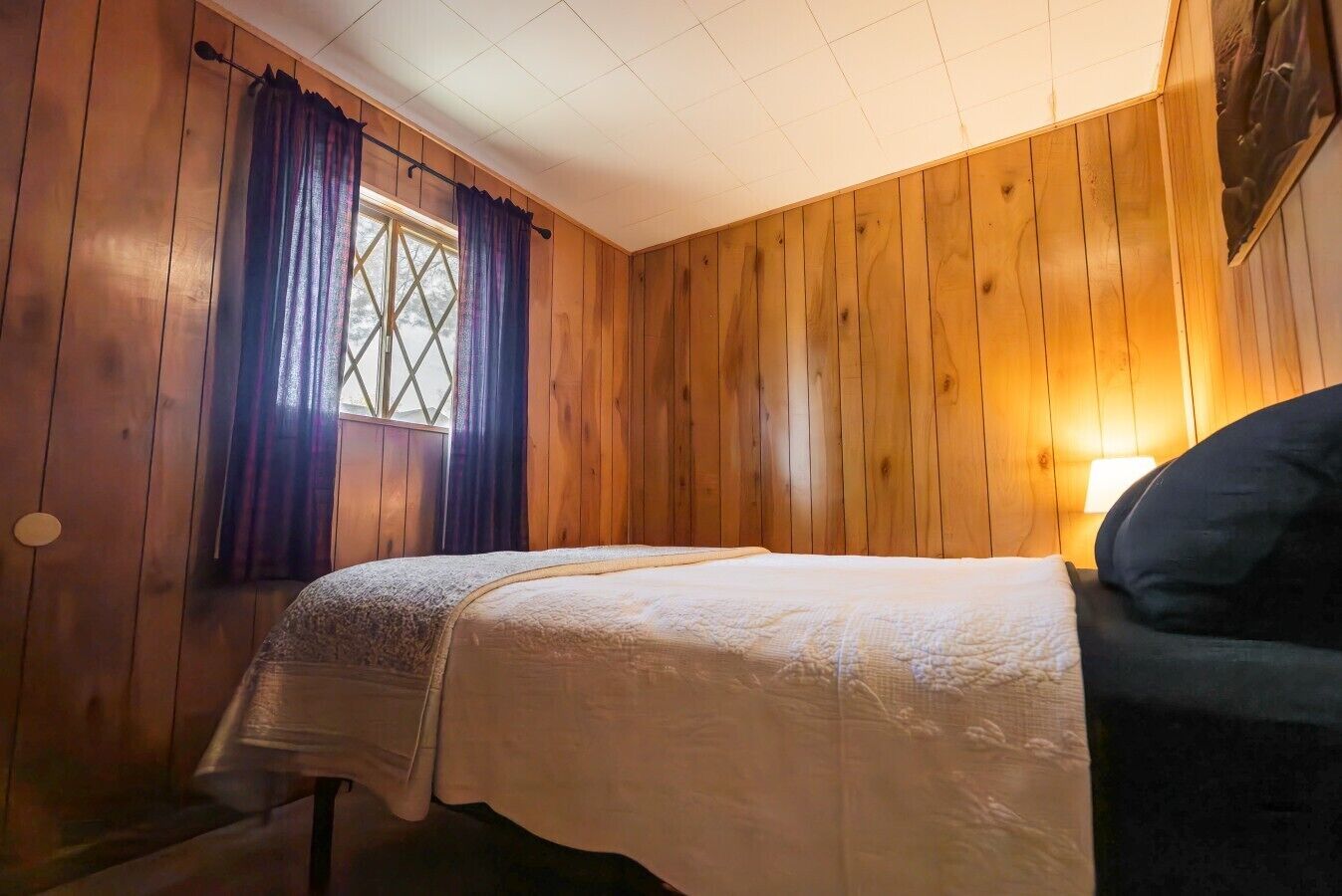 Quarto com paredes de madeira, cama com colcha branca, janela com cortinas roxas e luminária fixada na parede.