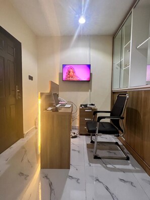 A reception area with a wooden desk, a black office chair, a television mounted on the wall, and a marble-tiled floor. - All States Apartments (Abuja)