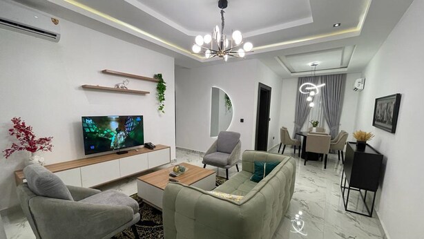 A modern living room with a flat-screen TV, a sofa, two armchairs, a coffee table, and a dining area with a table and chairs. - All States Apartments (Abuja)