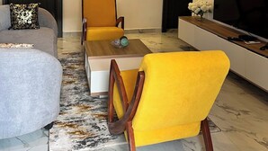 A modern living room with a sofa, two yellow chairs, a coffee table, a TV, and a unique ceiling light fixture. - All States Apartments (Abuja)
